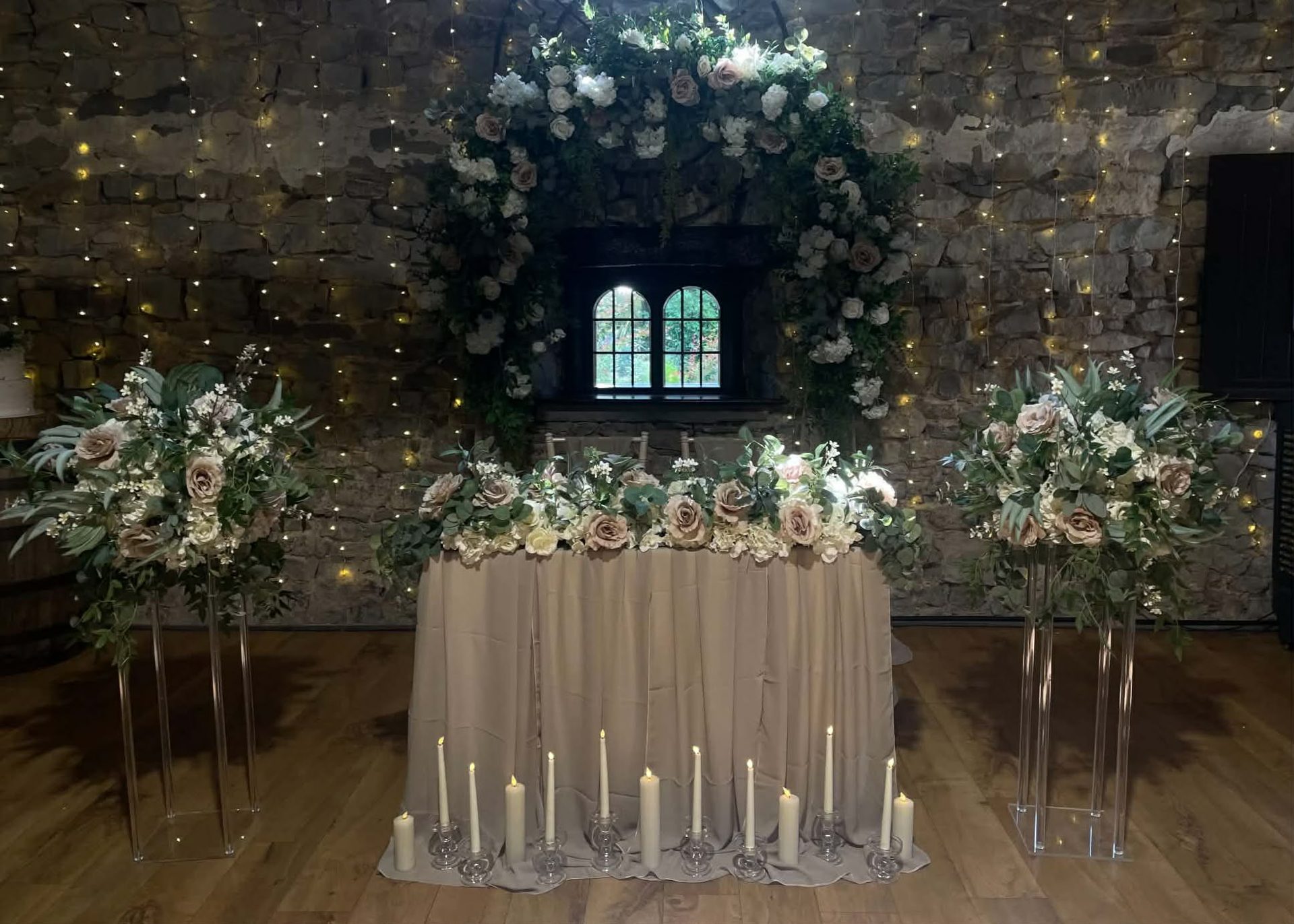 A sweetheart table beneath a blush and ivory floral arch against a stone wall strung with fairy lights, tall floral stands either side and taper candles along the floor.